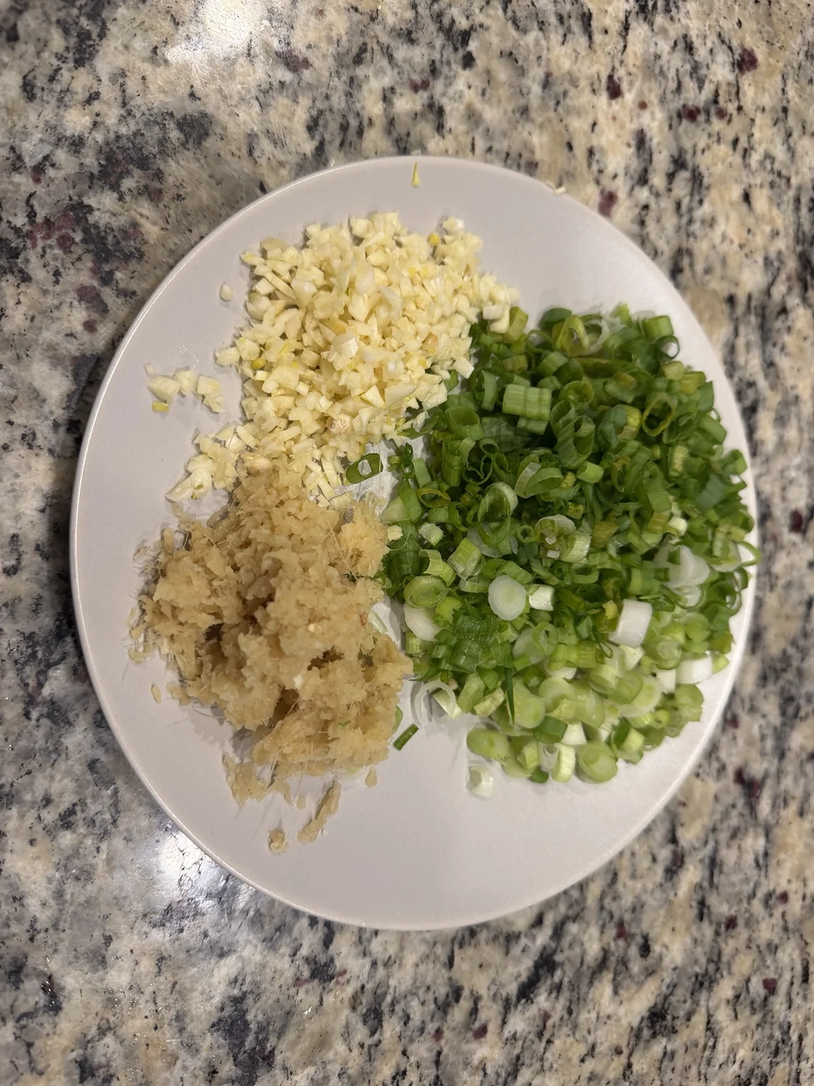A plate with freshly grated ginger, minced garlic, and chopped green onions in separate piles.