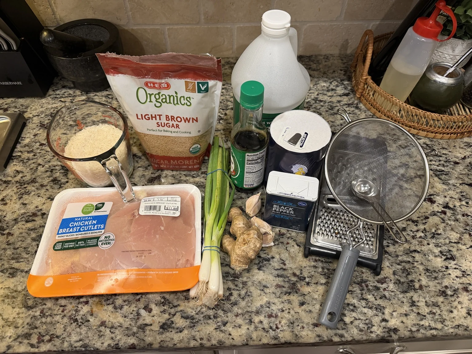 Raw chicken cutlets, jasmine rice, brown sugar, soy sauce, garlic, ginger, green onions, and kitchen tools arranged on a countertop.