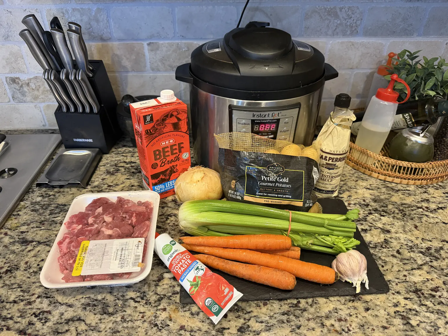 Beef stew meat, petite gold potatoes, carrots, celery, onion, garlic, beef broth, tomato paste, and Worcestershire sauce on a countertop in front of an Instant Pot.