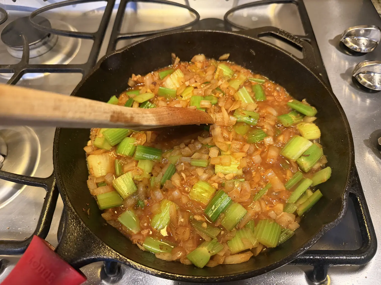 Onion, celery, garlic, and tomato paste in a cast-iron skillet being stirred with a wooden spoon.